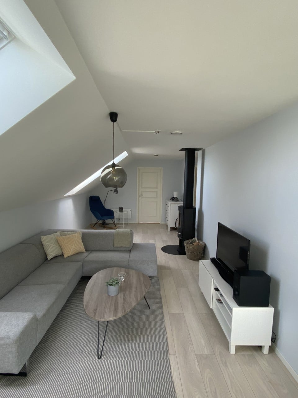 A light-filled living area features a modern, L-shaped gray sofa arranged around a small wooden coffee table. A television sits on a white media unit. A small black wood-burning stove is positioned against a pale wall, complemented by an overhead pendant light.