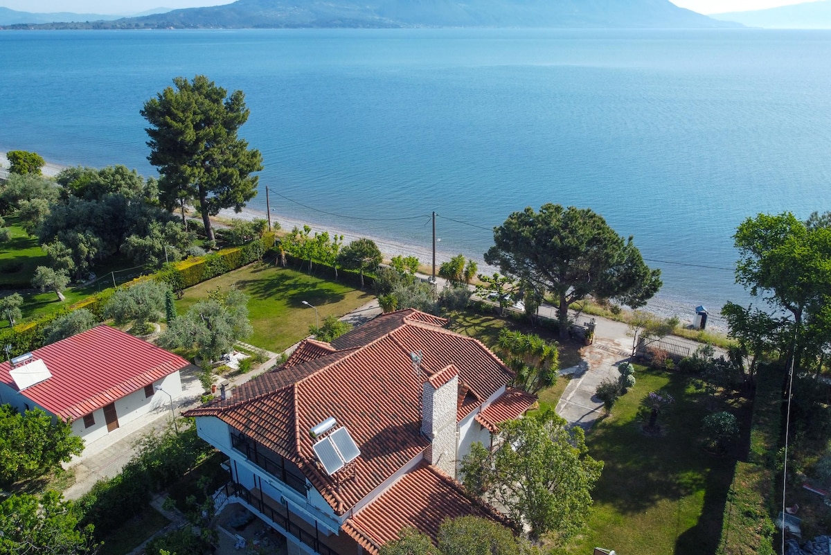 An aerial view reveals the beachfront apartment surrounded by greenery and a well-maintained garden. The red-tiled roof complements the coastal scenery, with a sandy beach visible nearby. A tranquil sea extends beyond, offering a serene backdrop to the property.