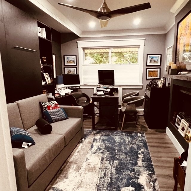 A functional office space is shown, featuring a gray sofa and a desk with a computer setup. Framed photos adorn the walls, while a blue and white area rug adds a touch of color to the wood floor. A ceiling fan offers ventilation.