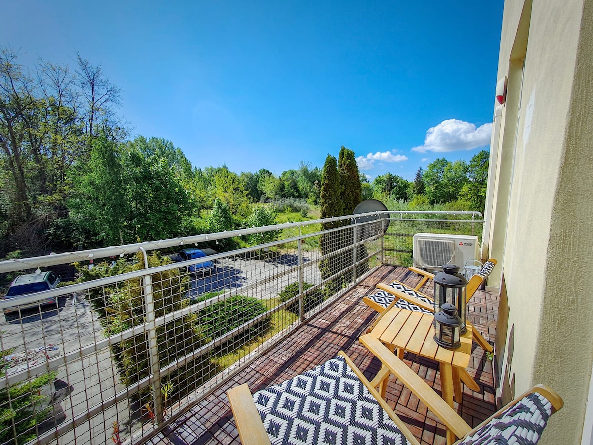 A private balcony features two wooden chairs and a small lantern, overlooking a green landscape with trees. An air conditioning unit is visible, and a parking area with cars can be seen below. The sky is clear with a few clouds, creating a bright atmosphere.