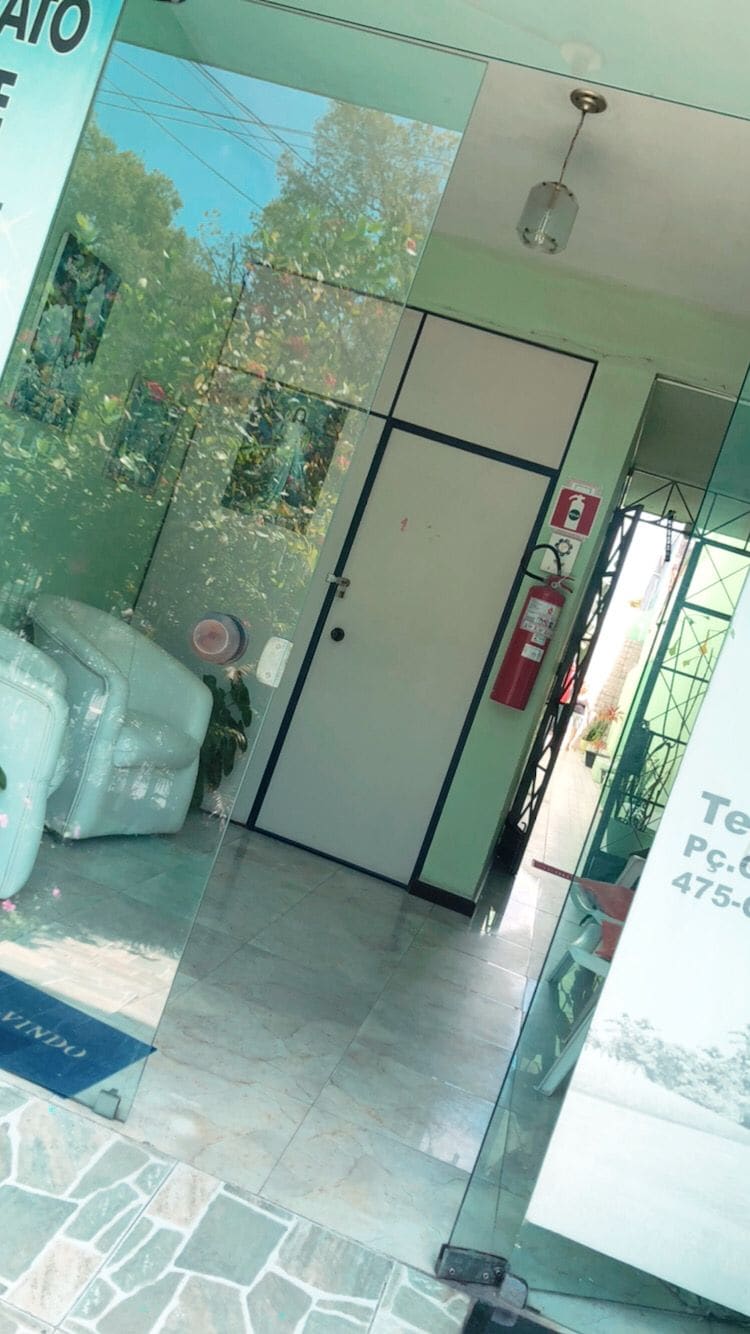 A bright entryway presents a mirrored glass door leading to interior spaces. Green plants are visible, contributing to a fresh ambiance. Light-colored tiled flooring enhances the openness of the area, while a fire extinguisher is positioned for safety.