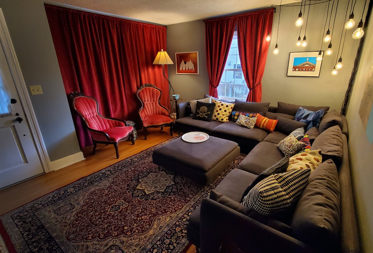 A cozy living area features a spacious sectional sofa adorned with various decorative pillows. Two antique-style chairs are positioned nearby, and warm light is provided by a standing lamp. The room is enhanced by rich red curtains and a patterned area rug, creating a comfortable ambiance.