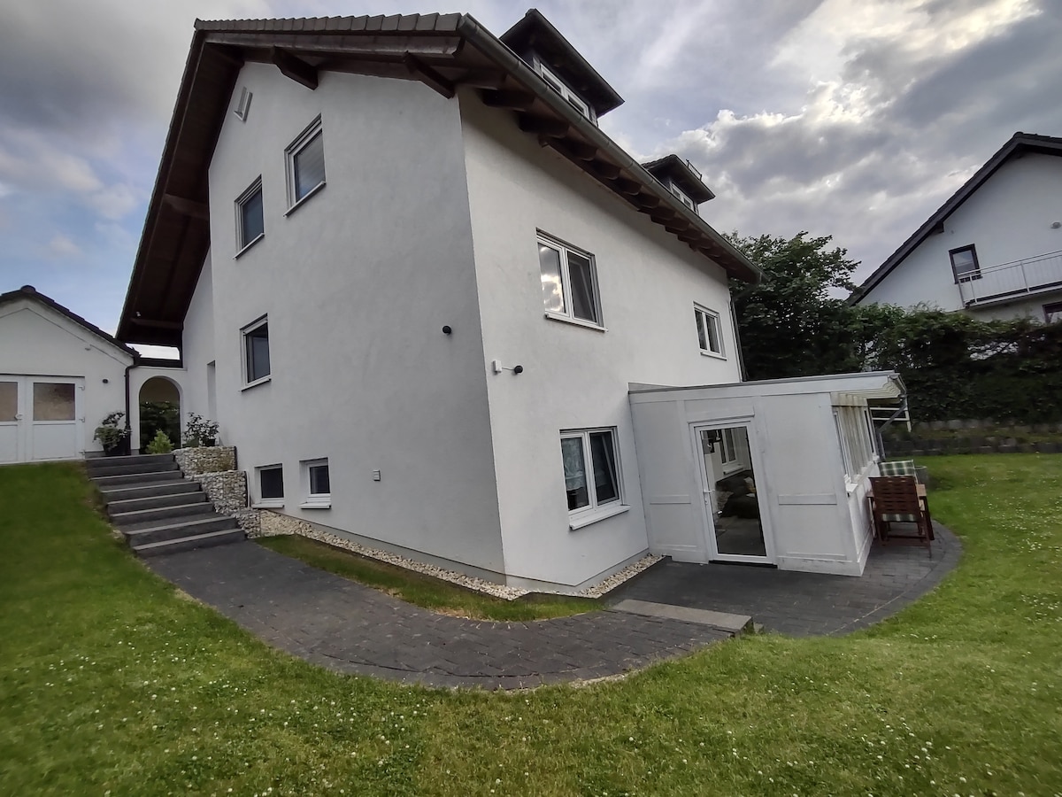 The exterior of a two-story house is shown, featuring a white facade and a sloped roof. A pathway leads to a winter garden with large windows, alongside a well-maintained lawn. Surrounding greenery enhances the tranquil setting.