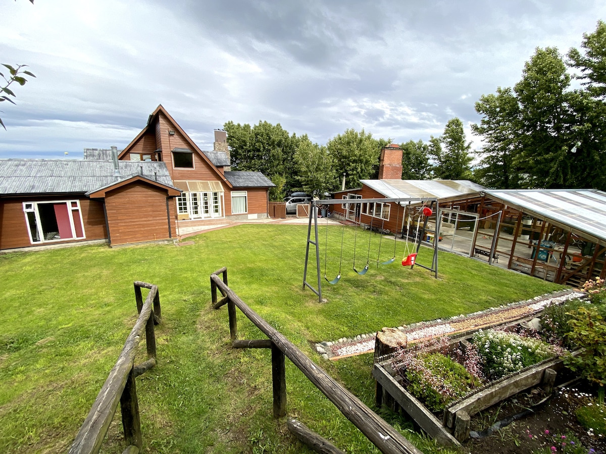 A spacious outdoor area is visible, featuring a well-maintained lawn with a swing set. The wooden house is situated in the background, complemented by a covered patio space. Surrounding greenery creates a peaceful environment, ideal for relaxation and gatherings.