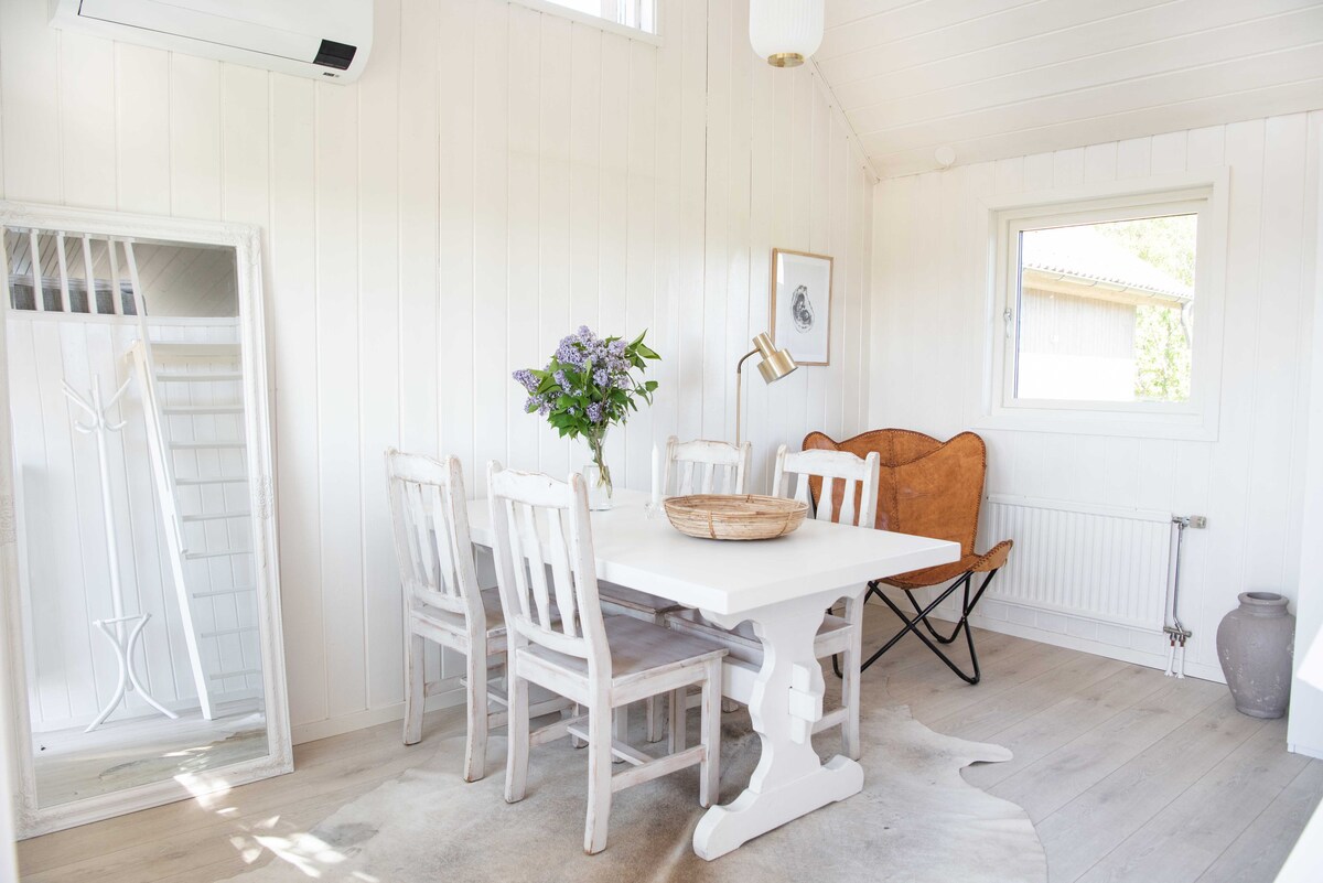 A light-filled dining area features a white table surrounded by six wooden chairs. A bouquet of flowers sits at the center of the table. A vintage mirror leans against the wall, and a cozy, tan armchair is positioned nearby, completing the inviting space.