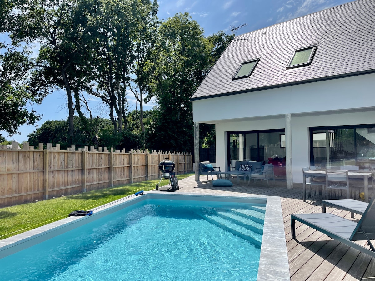 A view of the house and pool area is captured under clear skies. The heated swimming pool is centered within a wooden deck, surrounded by lounge chairs. Lush greenery and a wooden fence provide privacy, while large windows of the house invite natural light into the interior.