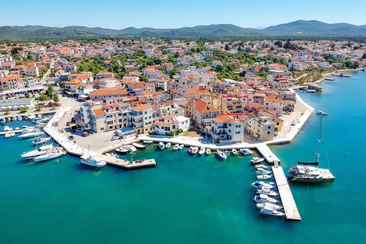 An aerial view showcases a coastal town with colorful buildings lining the waterfront. A variety of boats are docked along a pier, while the clear blue water reflects the sun. Green hills form a backdrop, enhancing the scenic coastal landscape.