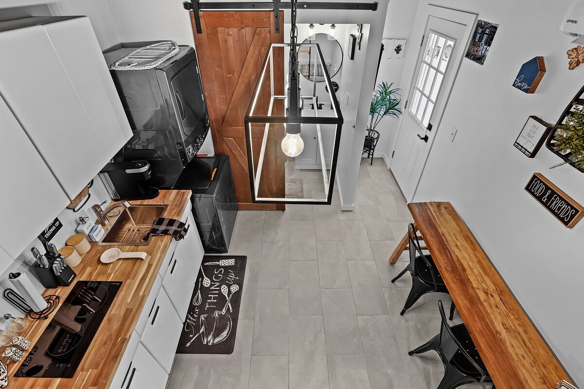 An airy kitchen space features white cabinetry, a wooden dining table, and sleek black chairs. A modern light fixture hangs above the table, and essential kitchen appliances are neatly organized. The room is well-lit by natural light entering through a nearby window.