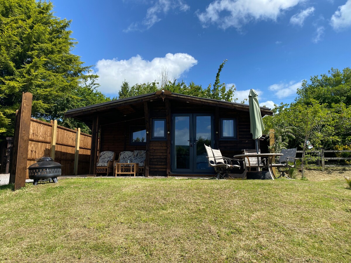 A wooden lodge is surrounded by greenery, featuring large glass doors that invite natural light. Outdoor seating is arranged on a grassy area, complemented by an umbrella and a fire pit nearby. The structure offers a rustic yet welcoming presence in a peaceful setting.