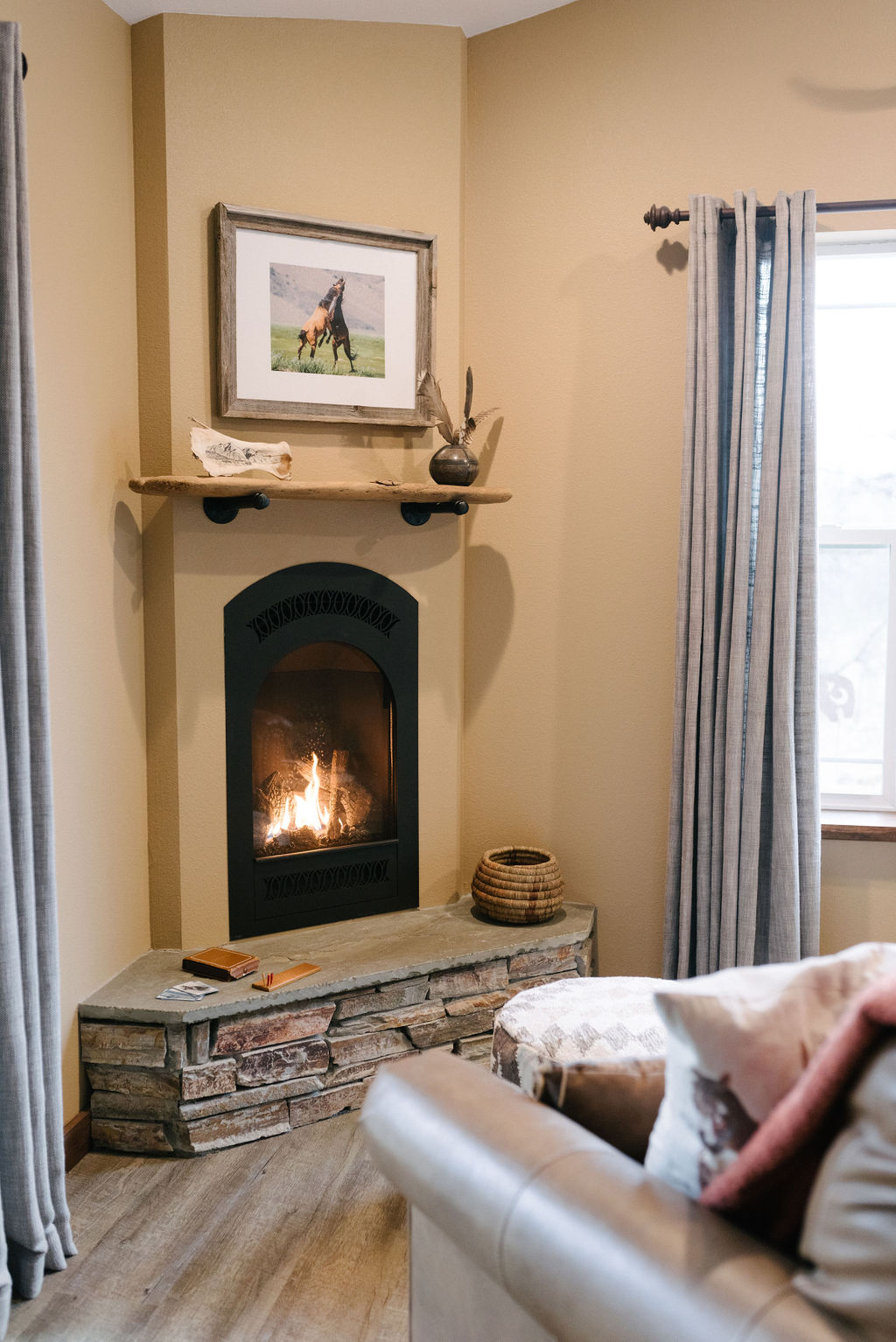 A cozy sitting area is highlighted by a gas fireplace, set within a stone surround. Above the fireplace, a framed artwork of a horse adds character. Soft gray curtains frame the window, allowing natural light to filter into the space.