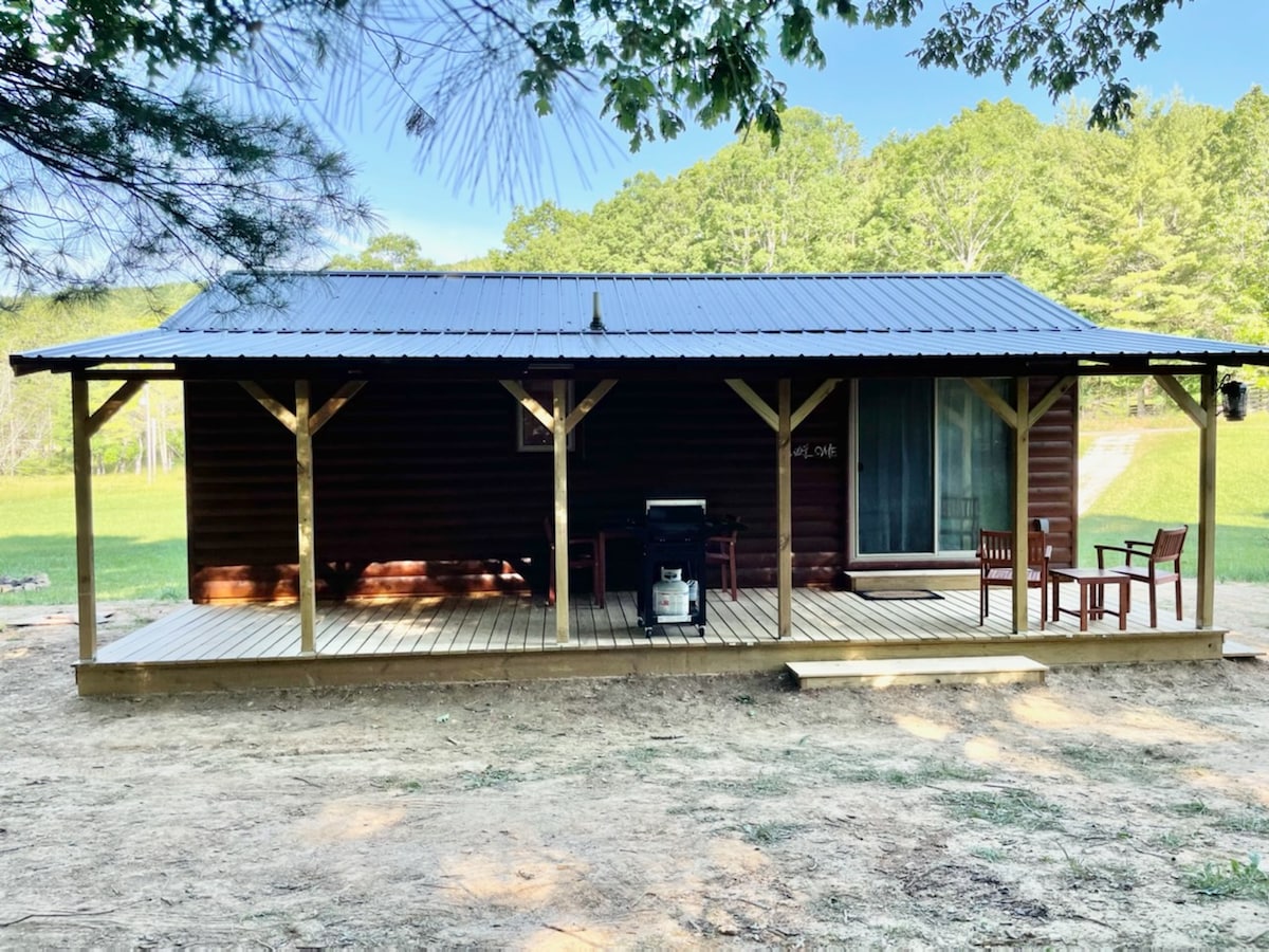 A rustic cabin is shown against a backdrop of lush greenery. The porch features wooden posts and a deck with seating arrangements, including four chairs and a grill. Large windows allow for natural light, enhancing the inviting exterior of the cabin.
