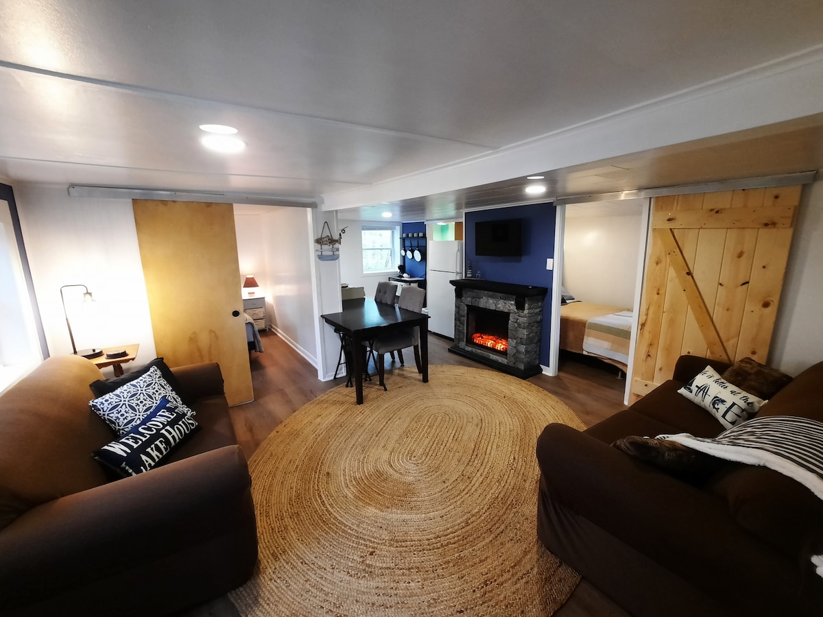 A cozy living area features two brown couches and a circular jute rug. A small dining table is set nearby, accompanied by a fireplace that adds warmth to the space. Soft lighting illuminates the area, enhancing the inviting atmosphere.