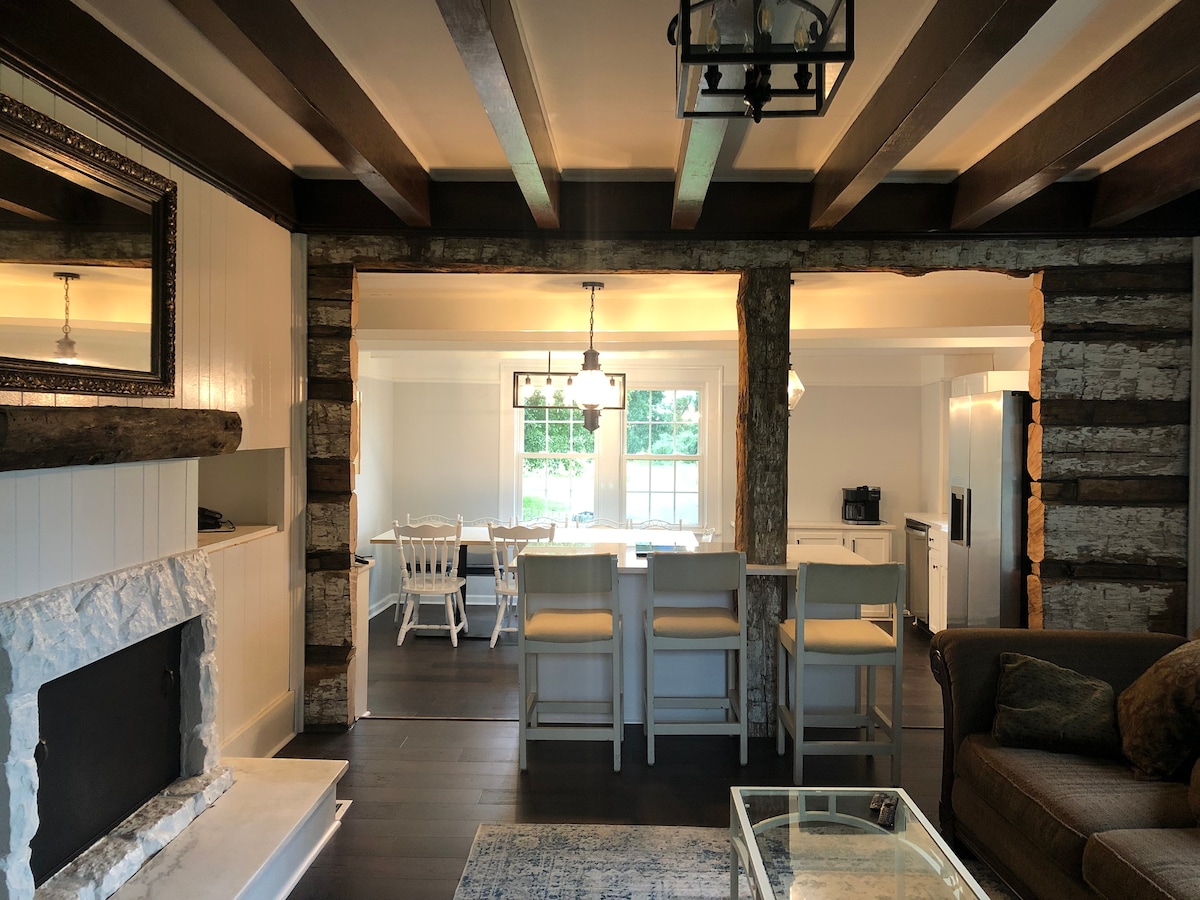 An inviting living area features wooden beams and a stone fireplace. A comfortable sofa contrasts with a glass coffee table. The adjoining dining space is visible, showcasing a large window that allows natural light to illuminate the white dining table and chairs.