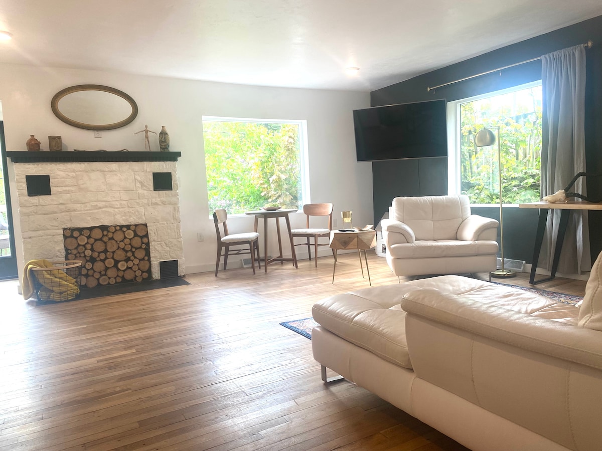 A spacious living room features a white stone fireplace surrounded by stacked wood. Large windows offer natural light and views of greenery. Modern furniture includes a comfortable white sofa, two chairs, and a small dining table, creating an inviting atmosphere for relaxation.