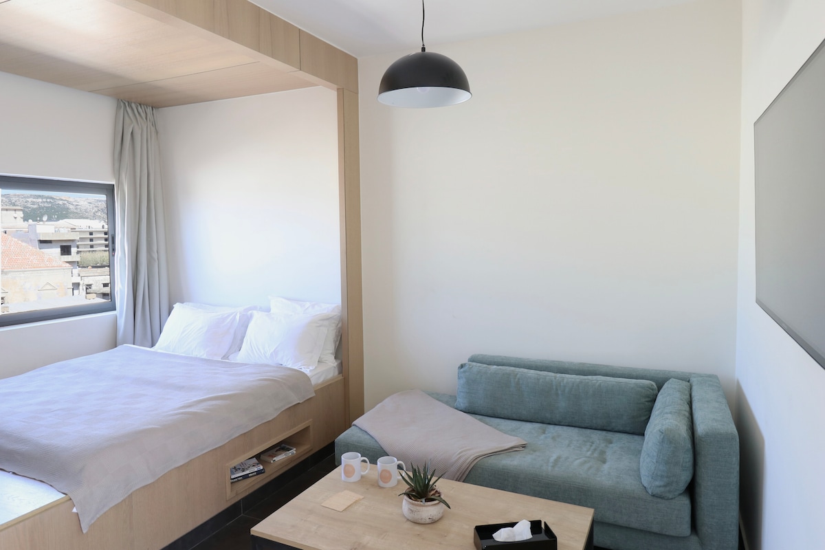 A bright studio space features a cozy sleeping area with a large bed and neatly arranged linens. A modern sofa sits beside a wooden table, accompanied by a small plant. Natural light enters through the window, creating a serene ambiance.