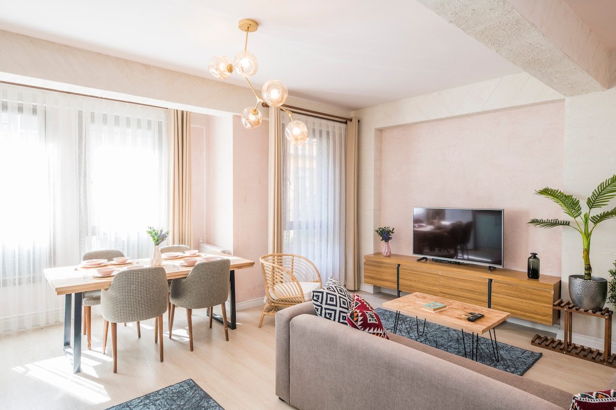 A modern living area features a light-toned sofa and two complementary coffee tables. A dining table with four chairs stands adjacent, illuminated by a stylish chandelier. A television is positioned on a wooden console, with potted plants adding a touch of greenery to the space.