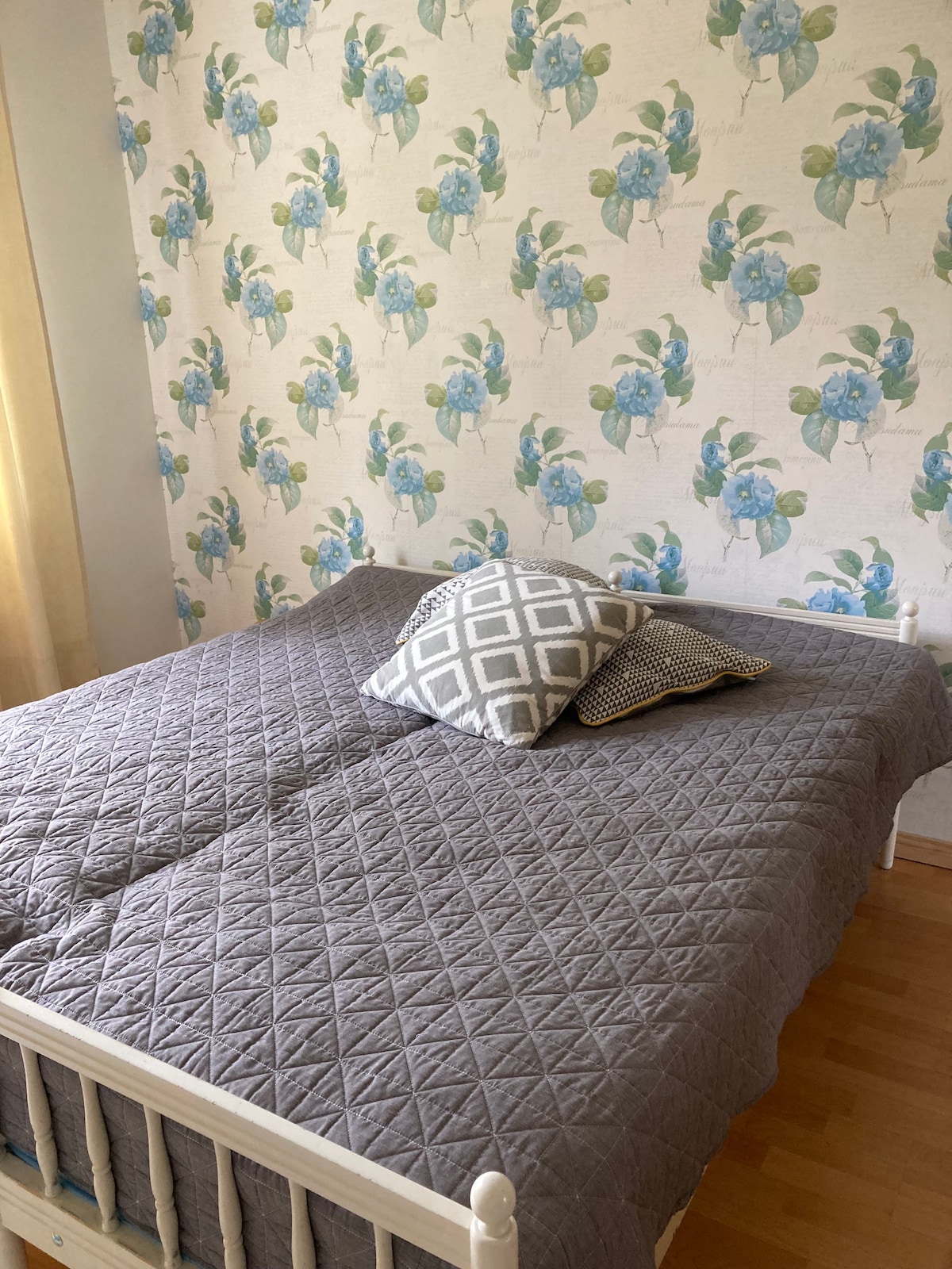 A sleeping area features a spacious bed covered with a grey quilt. Decorative pillows add texture. The walls display a floral wallpaper design in soft colors, while natural light filters through a nearby curtain, creating a calm ambiance.