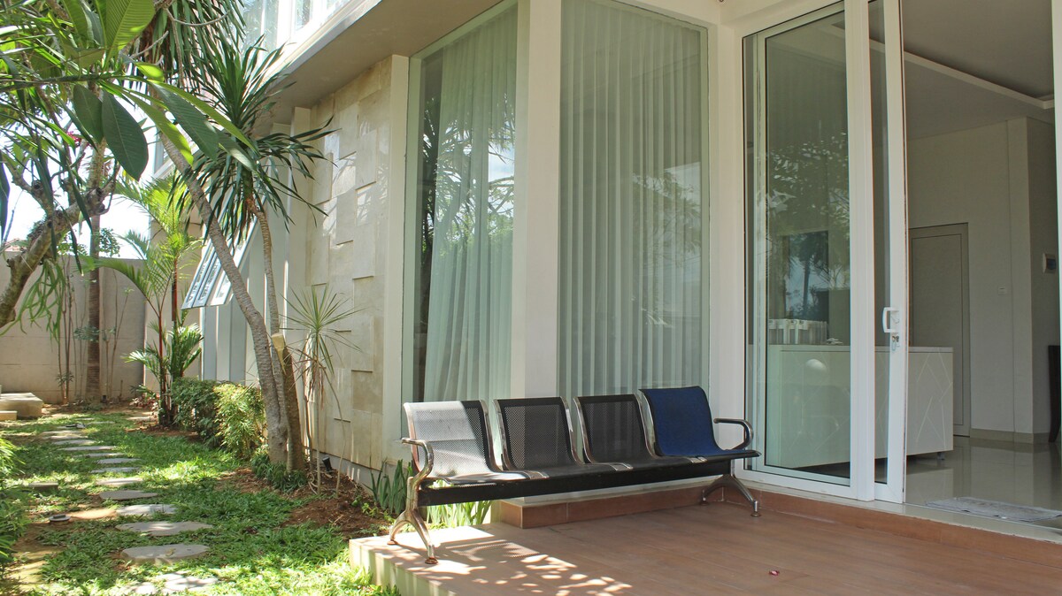 A sleek outdoor seating area features a modern bench against a backdrop of large windows. Lush greenery surrounds the patio, enhancing the relaxed atmosphere. The property layout showcases a blend of natural elements and contemporary design.