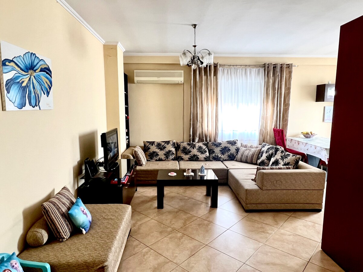 A spacious living room is furnished with a large sectional sofa decorated in a leaf pattern. A coffee table sits in the center, and a television is positioned on a nearby stand. Light pours in through sheer curtains, creating a bright and inviting environment.
