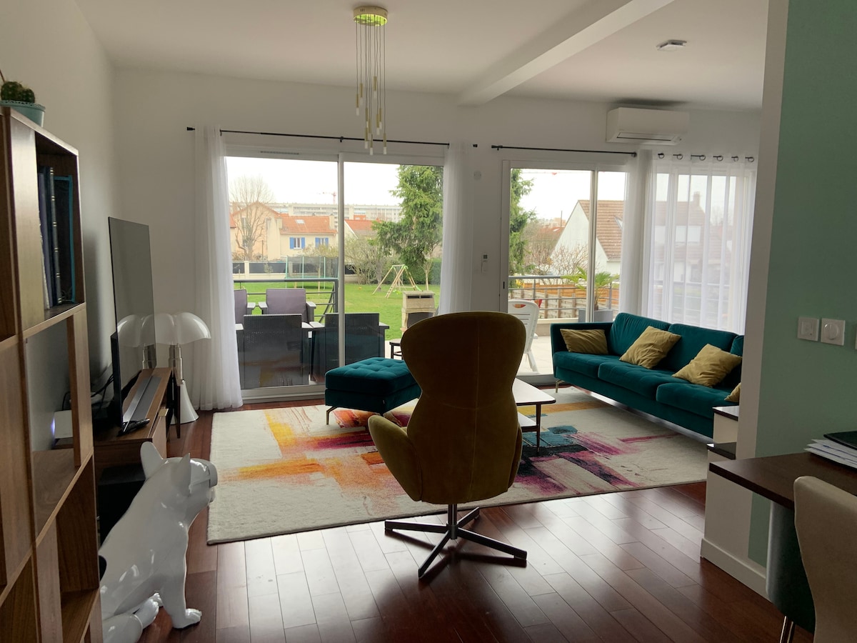 A welcoming living space features large glass doors that open to a garden view. A comfortable seating area includes a plush sofa and a vibrant accent chair, complemented by a colorful area rug. Natural light brightens the room, enhancing the modern decor.