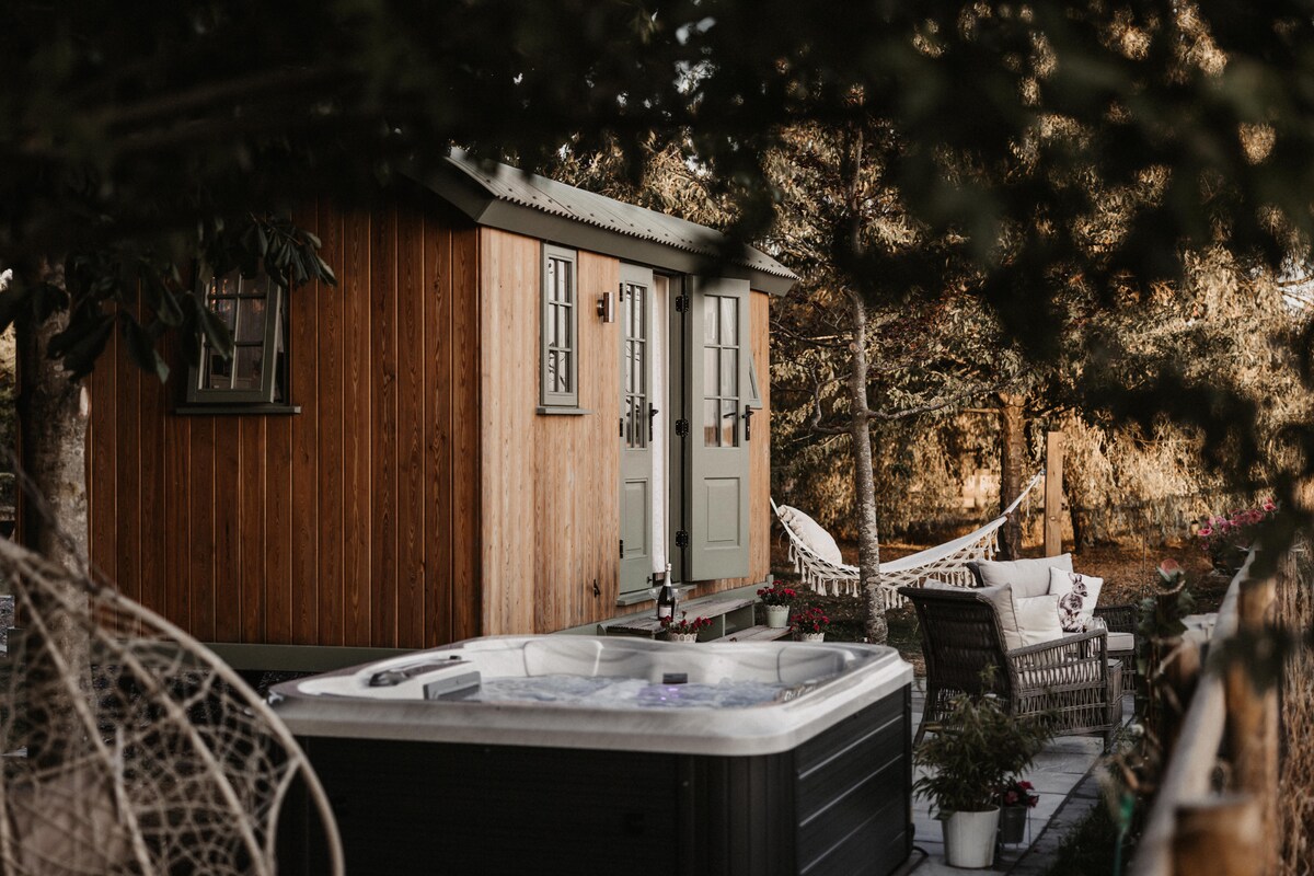A rustic wooden hut with double doors stands nestled among trees, complementing the tranquil surroundings. A luxury hot tub is positioned nearby, accompanied by a cozy seating arrangement. A hammock hangs in the background, inviting relaxation within a peaceful garden setting.