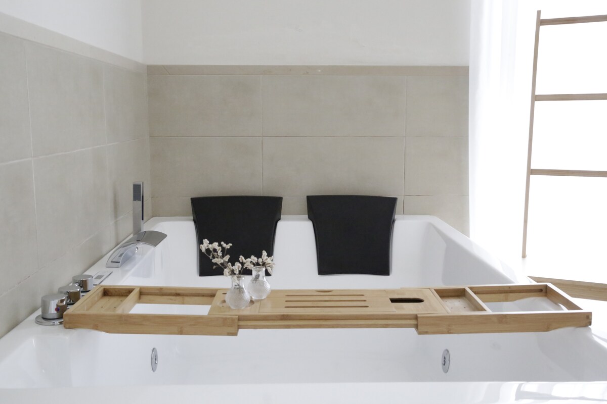 A spacious jacuzzi bathtub is featured, with a wooden bath tray placed across it. Two dark towels are elegantly draped over the sides. A small vase with flowers rests on the tray, enhancing the serene atmosphere of the bathroom.