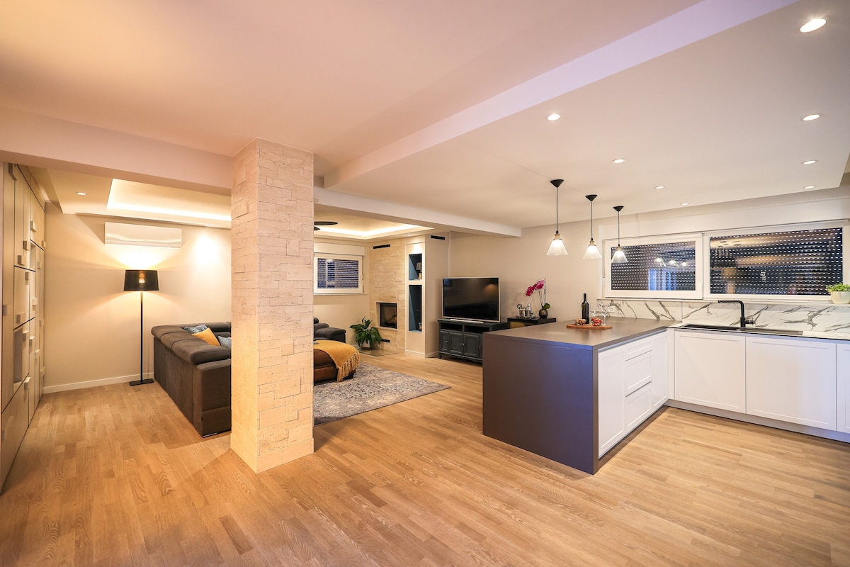 The open-concept living area features wooden flooring and modern decor. A spacious kitchen island is complemented by stylish pendant lighting. Adjacent, a comfortable sofa faces a flat-screen TV, with a cozy fireplace adding warmth to the space.