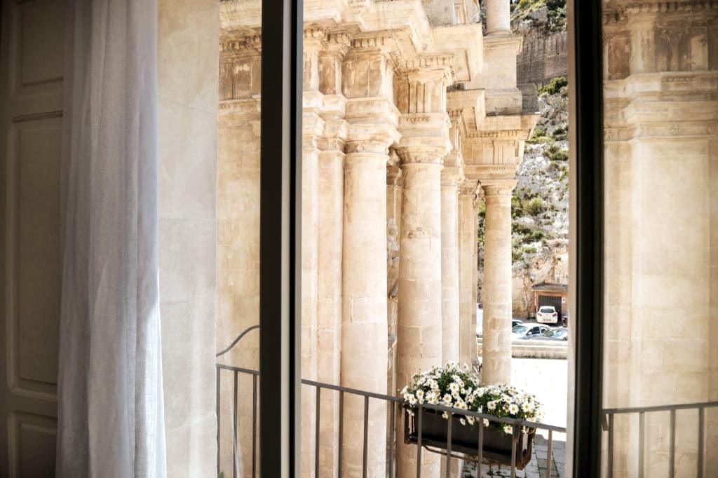 A view through large windows showcases grand columns of the Church of San Bartolomeo. A flower box filled with white blooms sits on the balcony railing, enhancing the connection between the indoor space and the historical architecture outside.