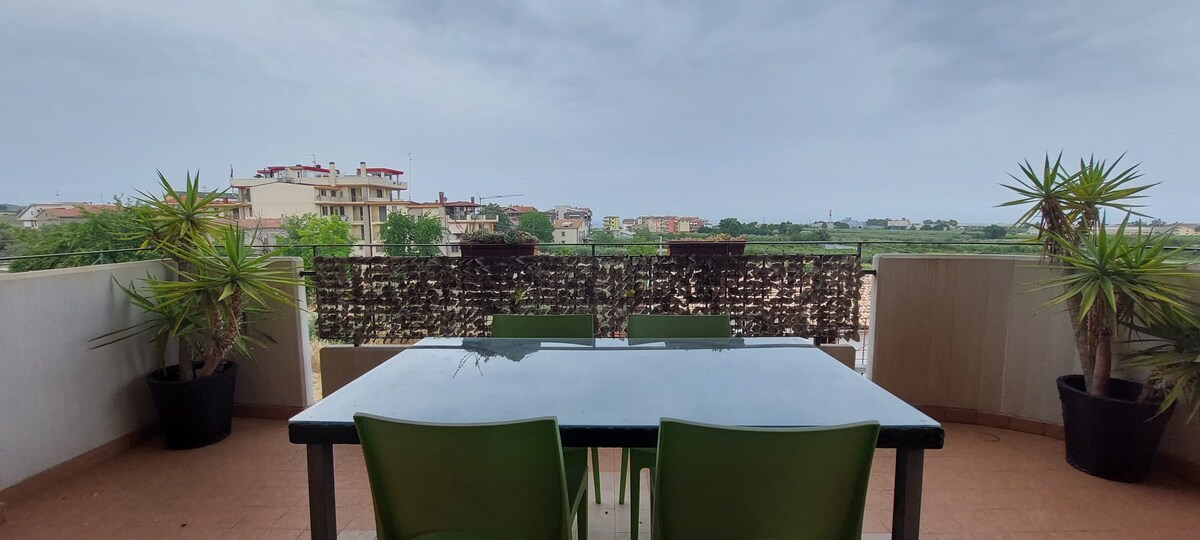 A balcony space features a sturdy black table surrounded by four green chairs. Potted plants are positioned on either side, offering a touch of greenery. The view includes residential buildings and an expansive landscape under a cloudy sky.