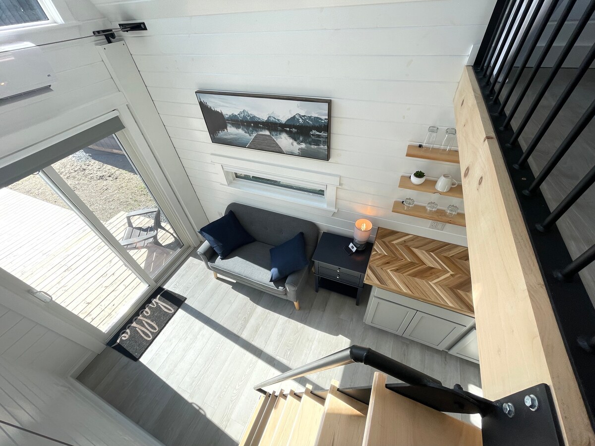 The interior of the tiny home is captured from an elevated angle, showcasing a cozy seating area with a blue sofa and a coffee table. Natural light streams through large sliding doors that lead to the outdoors, while a staircase and neatly arranged kitchen elements are visible.