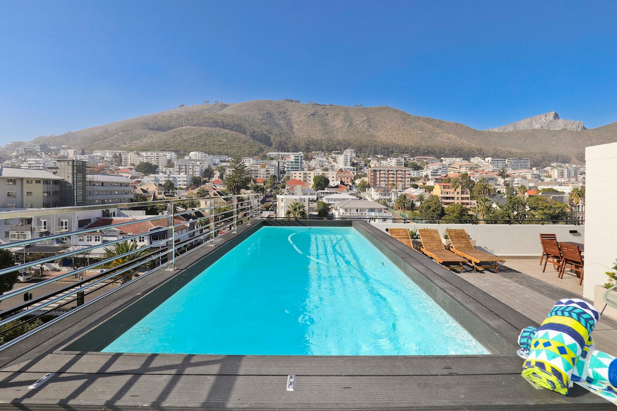 A rooftop swimming pool offers a serene escape, framed by a panoramic view of the surrounding hills and urban landscape. Sun loungers are positioned nearby, while colorful beach towels are neatly arranged at the pool's edge. Clear skies reflect in the inviting water.