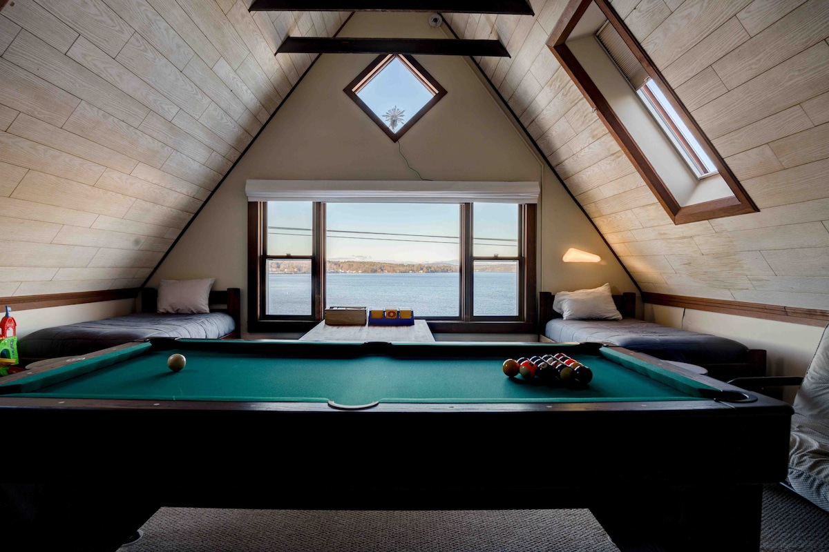 A triangular room features a pool table at its center, with two twin beds positioned against the walls. Large windows frame scenic views of the water beyond, allowing natural light to fill the space.