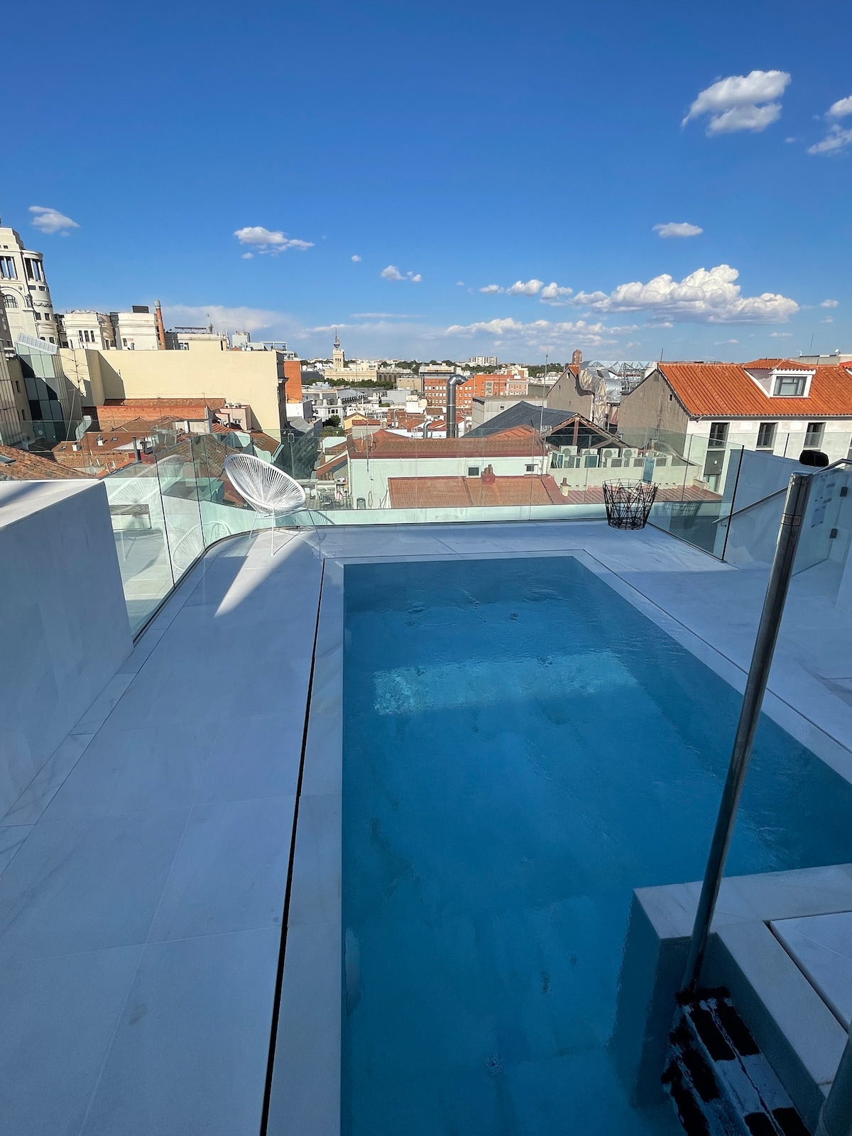 An outdoor terrace features a rectangular pool with clear blue water, surrounded by sleek glass panels. A panoramic view of the skyline, including rooftops and distant structures, is visible under a bright blue sky with scattered clouds.
