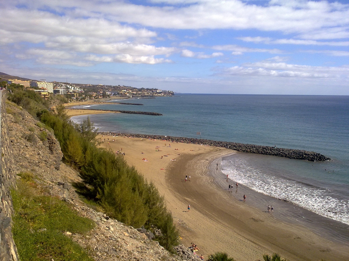 A sweeping view of a sandy beach is complemented by gentle waves lapping at the shore. A rocky jetty extends into the water, and distant buildings can be seen along the coastline under a sky dotted with clouds.