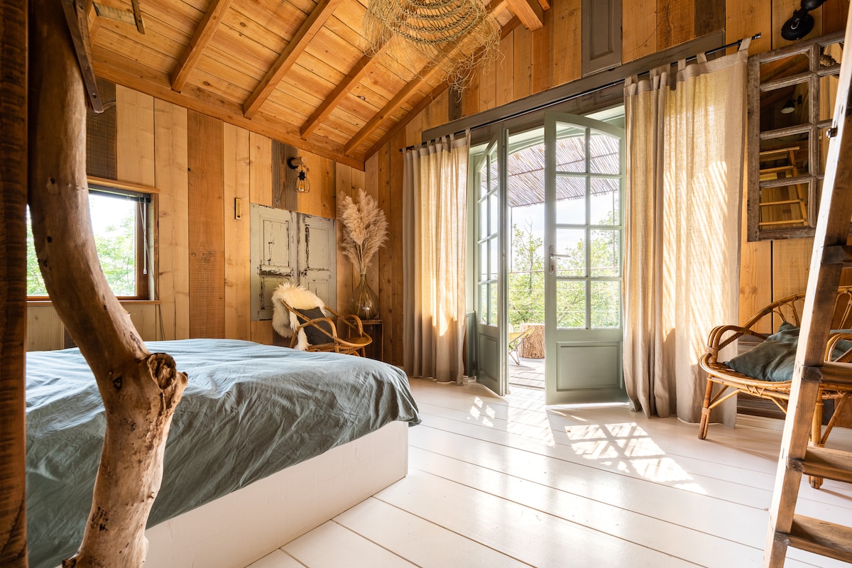 A bright interior features wooden walls and ceiling beams, creating a warm ambiance. A spacious bed with a soft blue cover is centered in the room. Large glass doors open to a terrace, complemented by flowing curtains and wooden furniture in the corners.