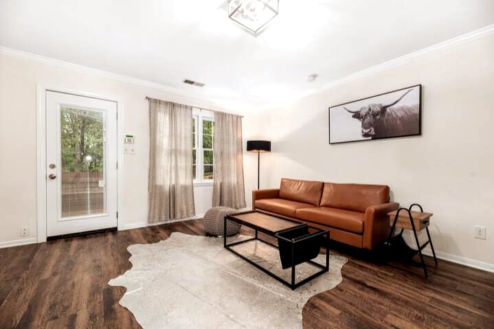A cozy living area features a leather sofa facing a coffee table, with a rug underneath. A framed artwork of a cow adorns the wall. Natural light enters through the sliding glass door, complemented by sheer curtains, creating a bright and inviting space.