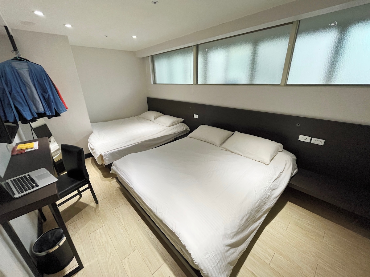 An en-suite room features two double beds positioned against a dark wooden wall. Natural light filters through frosted glass windows. A small workspace with a laptop and a chair is located beside a waste bin.