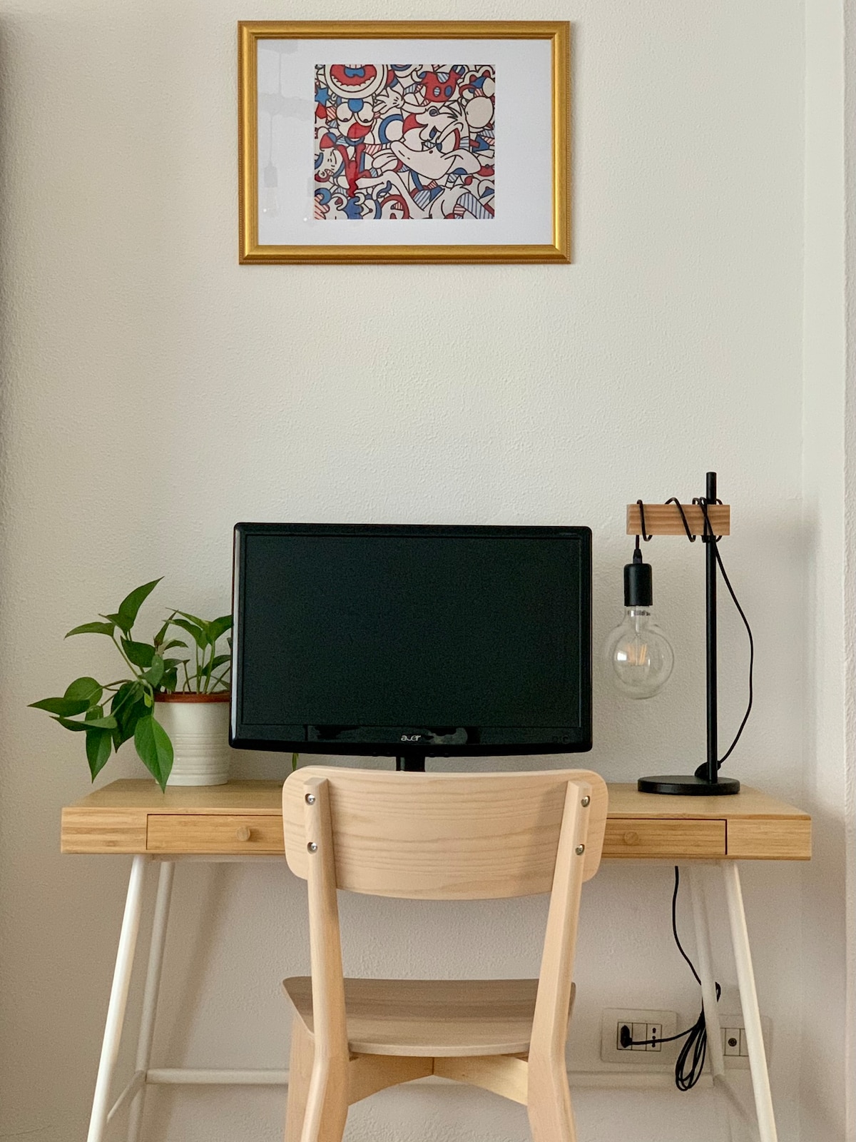 A simple workspace is highlighted by a wooden desk with a light-colored chair. A black monitor sits on the desk, accompanied by a modern lamp with a visible bulb. A potted plant adds a touch of greenery, while a framed artwork is displayed above.