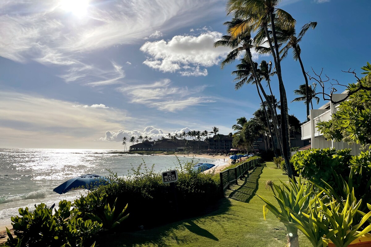 A scenic view showcases the beach and ocean, framed by palm trees and lush greenery. Umbrellas are positioned along the sand, while gentle waves reflect the sunlight. The horizon is dotted with buildings, enhancing the coastal atmosphere.
