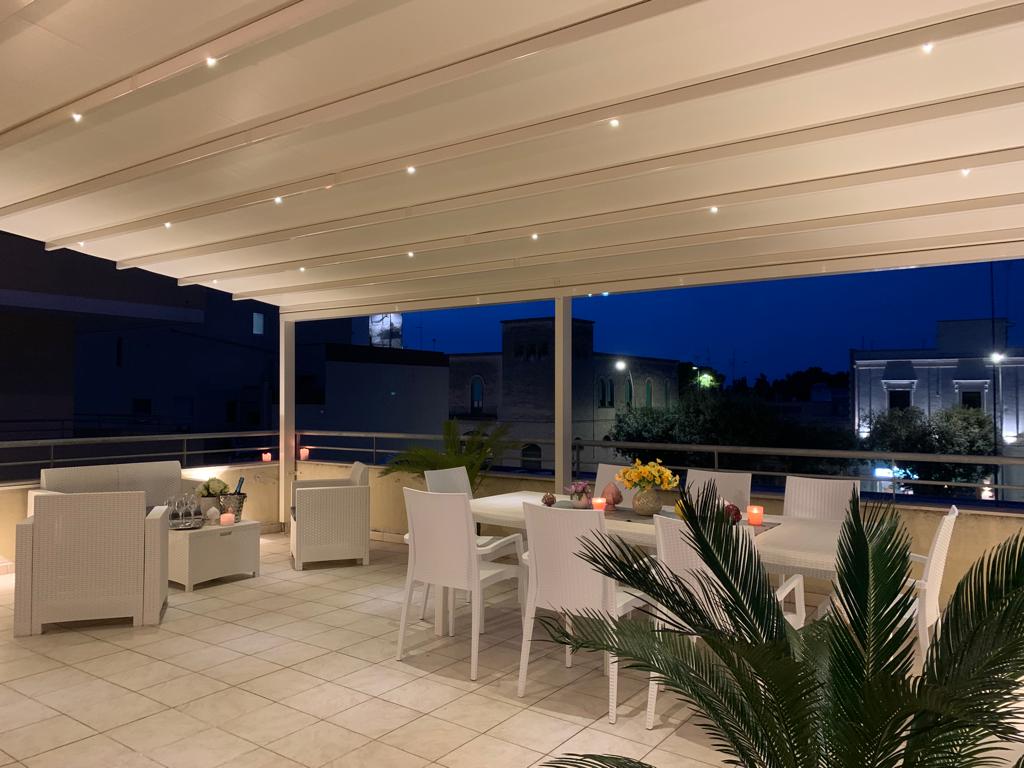 An outdoor terrace features ample dining space with a large table surrounded by eight chairs, adorned with decorative plants and centerpieces. The evening ambiance is enhanced by subtle lighting, creating a serene setting for relaxation. Surrounding buildings softly illuminate the night.