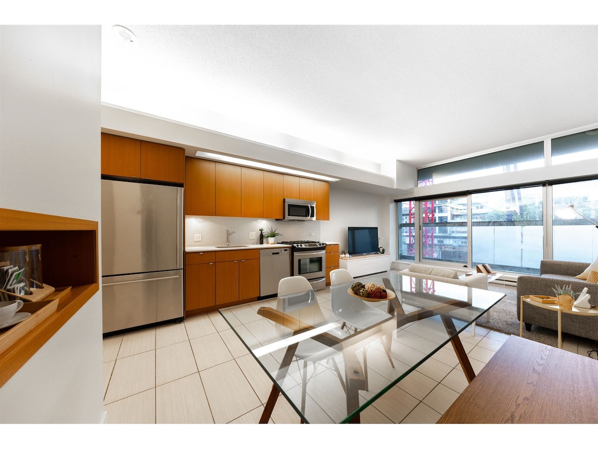 A modern kitchen features stainless steel appliances, including a refrigerator and oven. A sleek dining table is set with chairs, while natural light floods the space through large windows that offer a view. A comfortable sofa and a Smart TV complete the layout.