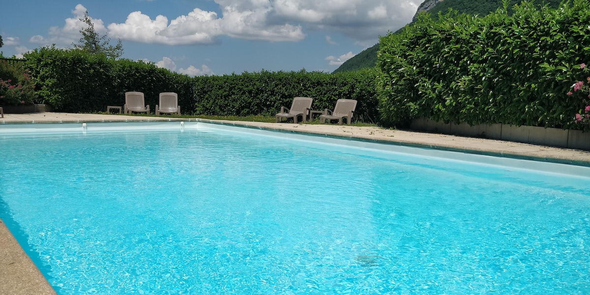 A clear blue swimming pool is framed by neatly trimmed hedges and sun loungers arranged around its perimeter. The surrounding landscape features lush greenery and distant mountains under a partly cloudy sky, creating a serene outdoor environment.