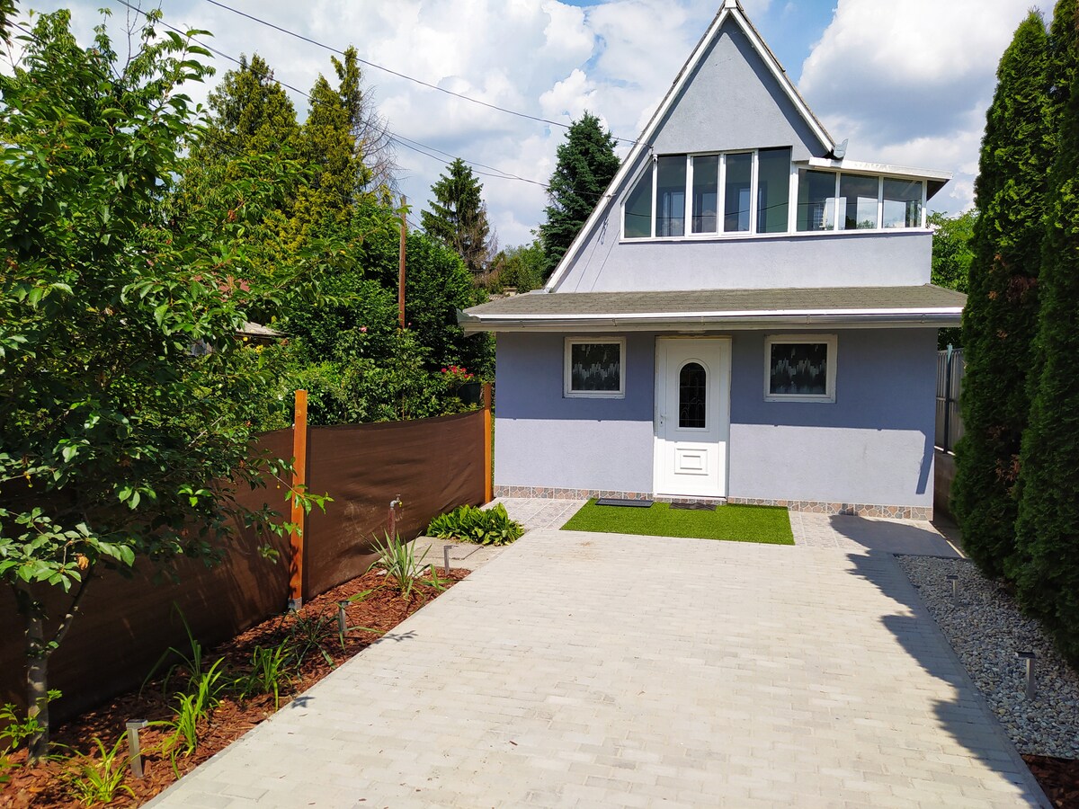 A charming view of the exterior features a two-story house with a sloped roof and a welcoming front door. The walkway is paved, bordered by landscaped areas with small plants and fresh greenery. A brown fence provides privacy, enhancing the peaceful setting.