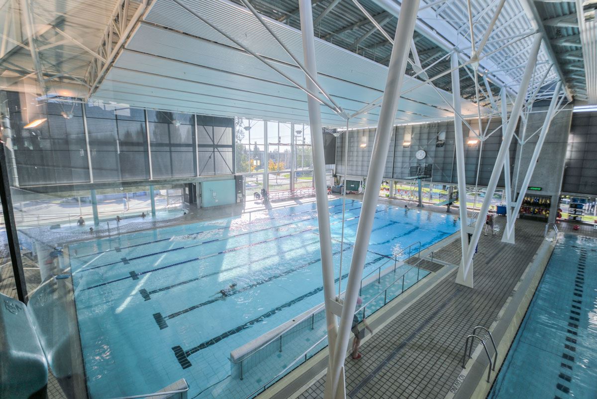 An indoor swimming facility is depicted, featuring a large pool with multiple lanes. The environment is brightened by natural light streaming through expansive windows, while spectators can be seen observing from a nearby seating area. Poolside amenities are also visible.