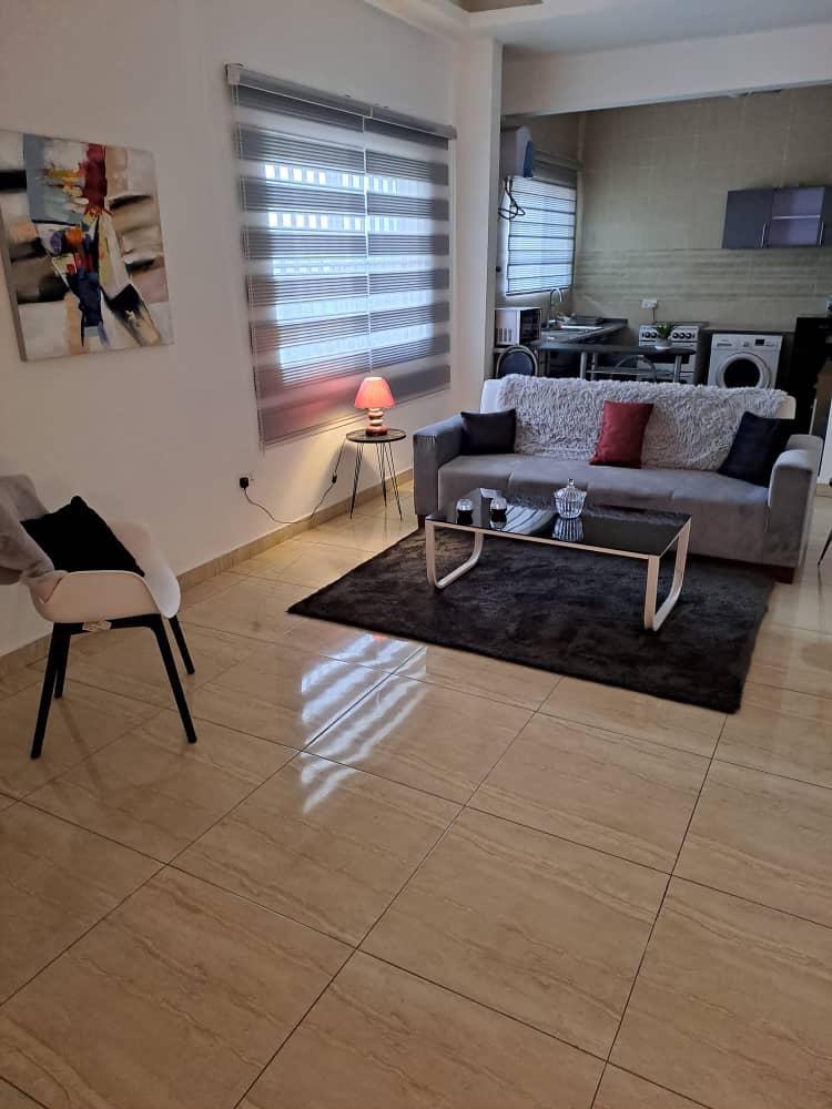 A modern living area features a light-colored sofa arranged on a plush rug, complemented by a glass coffee table. A small side table with a lamp provides soft lighting. A piece of abstract art adorns the wall, while sliding shades filter natural light from the windows.