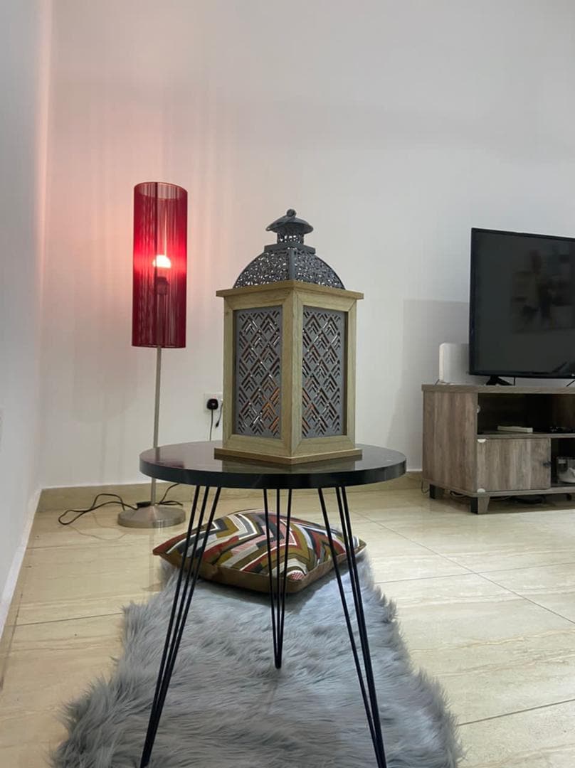 A decorative lantern is placed on a round black table, showcasing intricate designs. A vibrant red floor lamp stands nearby, providing soft illumination. A light-colored rug is positioned on the tiled floor, while a modern television unit can be seen in the background.