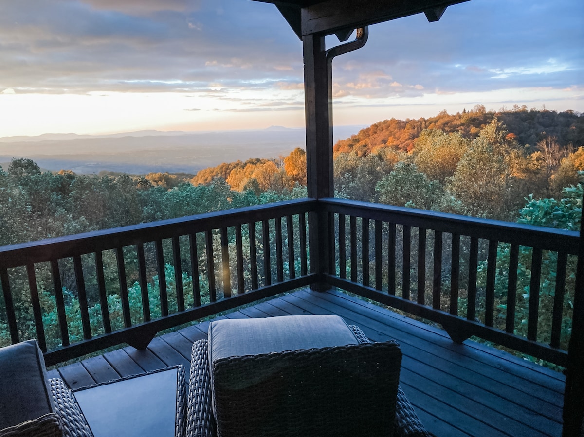 A deck presents a serene view of rolling hills and valleys bathed in soft morning light. Dark wooden railings frame the space, while two cushioned chairs invite relaxation. Lush greenery adorns the landscape, highlighting the beauty of the mountains beyond.