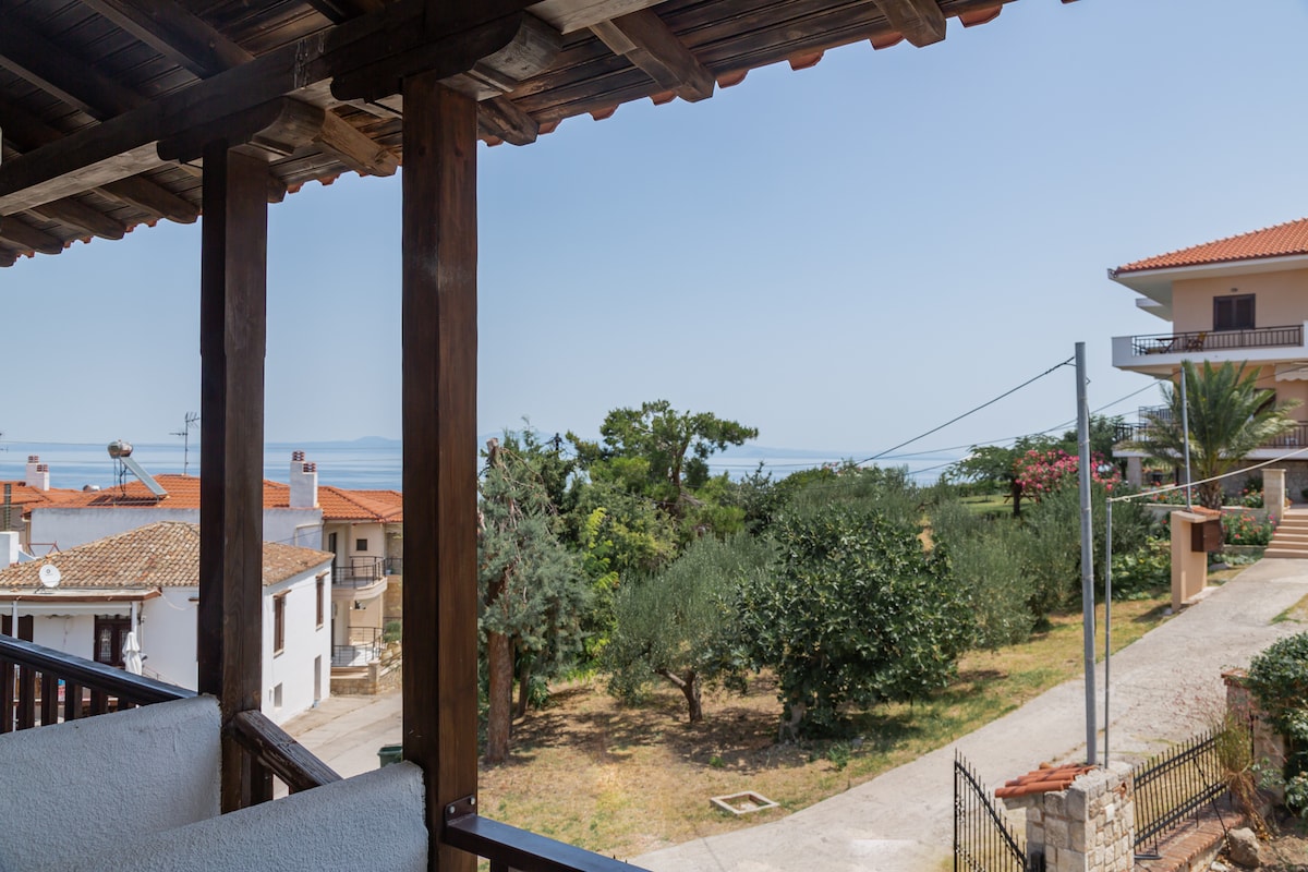 A view from the balcony reveals a serene landscape with greenery and a glimpse of the sea in the distance. The setting is enhanced by the wooden beams of the balcony, framing the scene and establishing a connection with the outdoors.