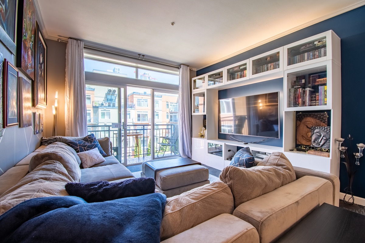 A spacious living area is shown, featuring a comfortable sectional sofa with varying fabric textures. A large television is mounted on a white entertainment unit, surrounded by shelves filled with books and decorative items. Oversized windows allow natural light to flood the room, offering a view of the outdoor balcony.