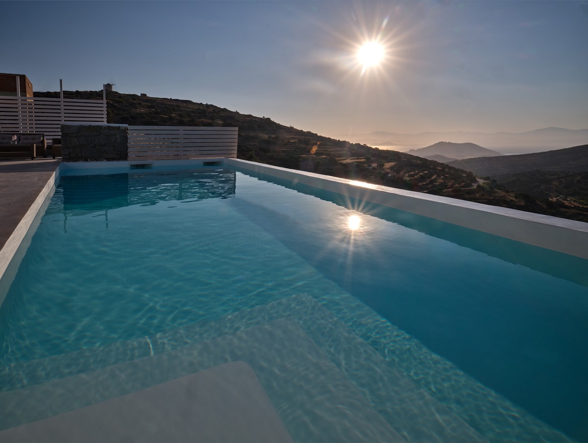 A serene private swimming pool is captured in the foreground, reflecting the sun's rays and the surrounding hills. The space is designed for relaxation, with smooth lines and a clear blue water surface, inviting tranquility and peace to guests.
